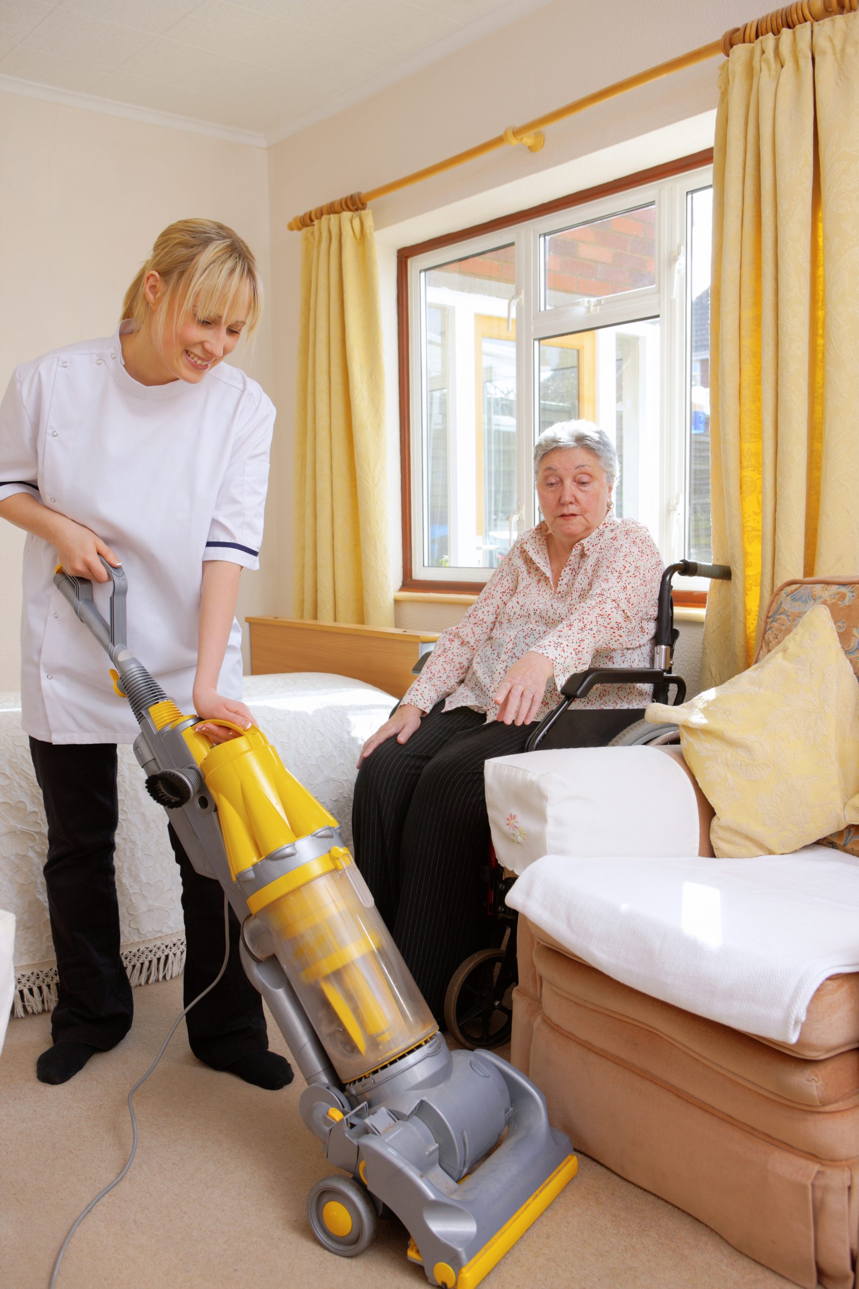 carer helps her elderly pacient by vacuuming her carpet for her